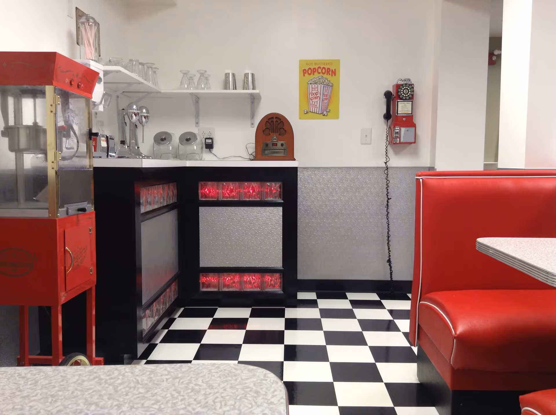 Interior of a retro-style diner area with black and white checkered floor, red cushioned booth seating, a red popcorn machine, a counter with glassware and vintage items, a wall-mounted red rotary phone, and a yellow popcorn sign on the wall.