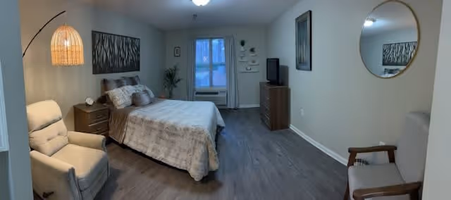 A senior living facility bedroom with a bed covered in a beige quilt, a wooden nightstand with a lamp, a beige armchair with a floor lamp beside it, a wooden dresser with a TV on top, a round mirror on the wall, and a window with curtains letting in natural light.