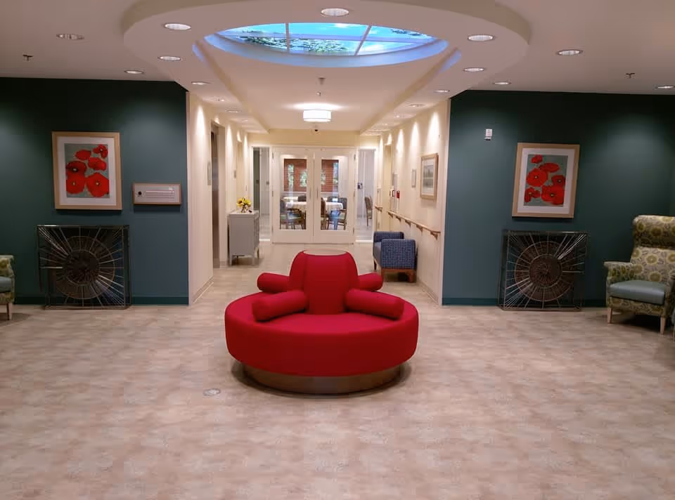 A spacious hallway in a senior living facility with a round red cushioned bench in the center. The walls are painted green and cream, decorated with framed pictures of red flowers. There are armchairs on either side of the hallway and a skylight above, allowing natural light to illuminate the area. At the end of the hallway, glass doors lead to another room with tables and chairs.