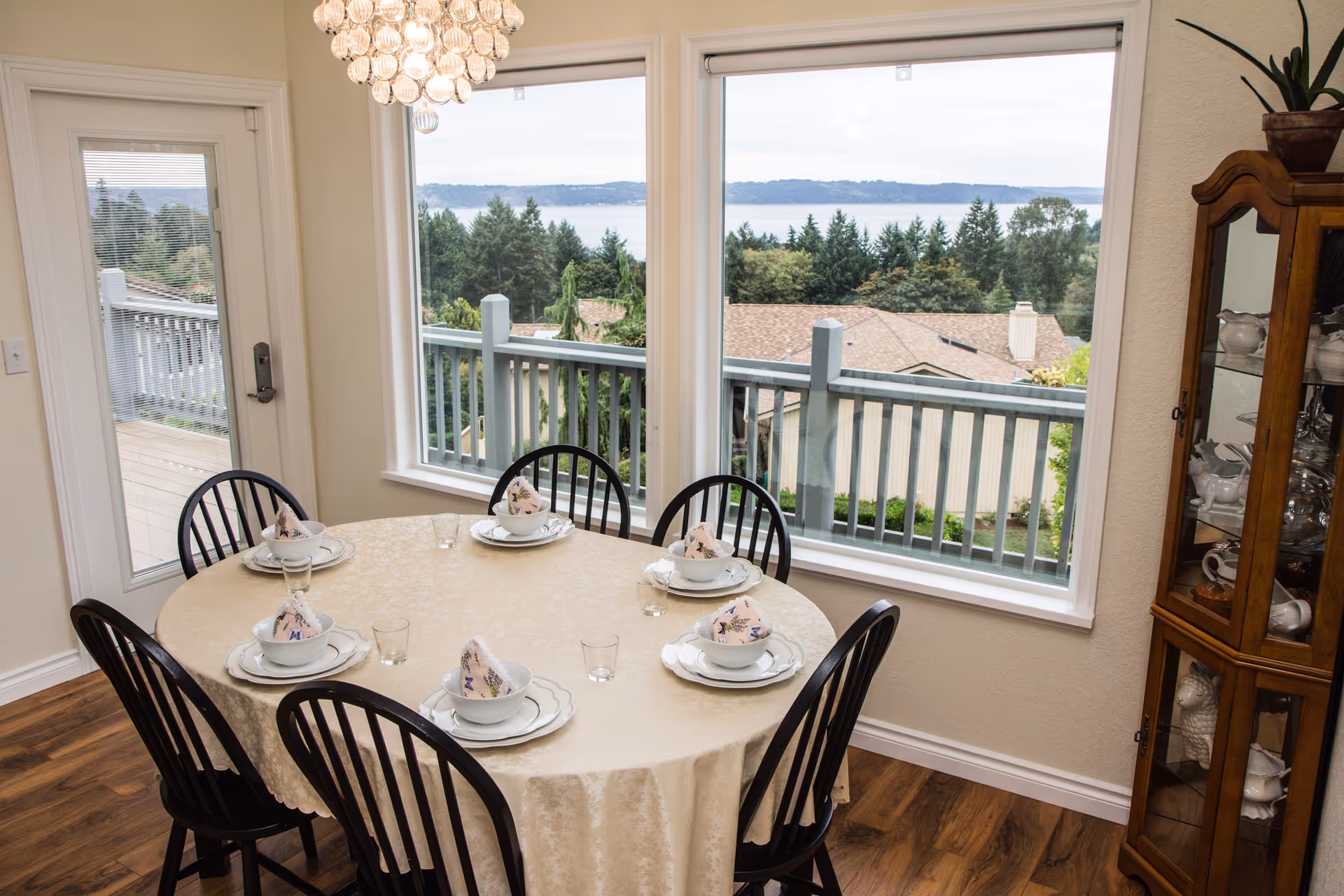 A dining room with a round table covered with a beige tablecloth set for six people with white plates, bowls, and floral napkins. The room has large windows offering a view of trees, rooftops, and a body of water in the distance. There is a glass door leading to an outdoor deck and a wooden cabinet displaying white dishware.