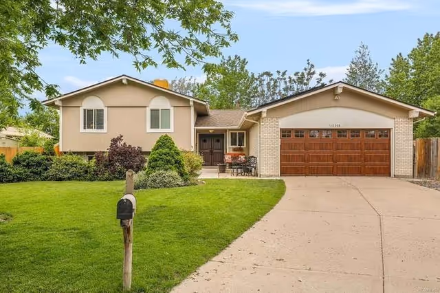 Exterior view of a single-story residential house with a well-maintained green lawn, a driveway leading to a wooden garage door, a mailbox on a post near the street, and some outdoor seating near the front entrance. Trees and shrubs surround the property under a clear blue sky.