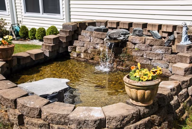 A small outdoor water feature with a stone brick wall and a spout pouring water into a shallow pond. There is a flower pot with yellow and orange flowers on the edge of the pond. The background shows a white building wall with windows and some green shrubs.