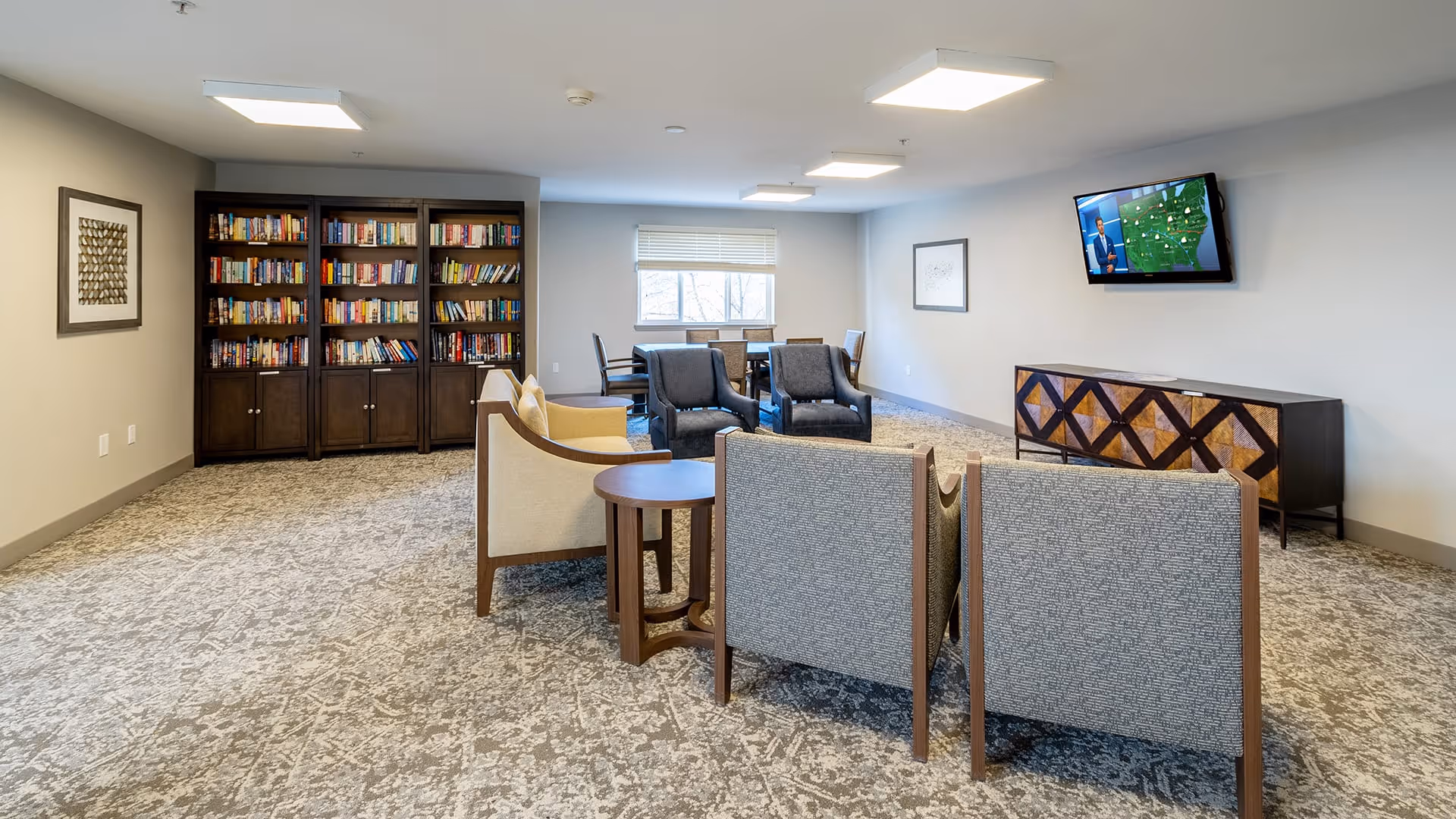 A spacious common area with multiple chairs arranged around small tables, a large bookshelf filled with books against the wall, a flat-screen TV mounted on the wall showing a weather forecast, and a sideboard with a geometric pattern. The room has carpeted flooring, neutral-colored walls, and a window letting in natural light.