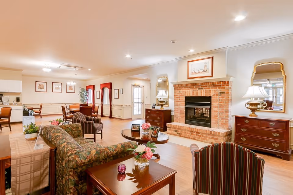 A cozy senior living common area featuring a brick fireplace with a framed picture above it, two wooden dressers with lamps and mirrors on either side, a floral patterned sofa, striped armchairs, and a round wooden coffee table with framed photos and flowers. In the background, there is a dining area with tables and chairs, wall art, and windows with red curtains.