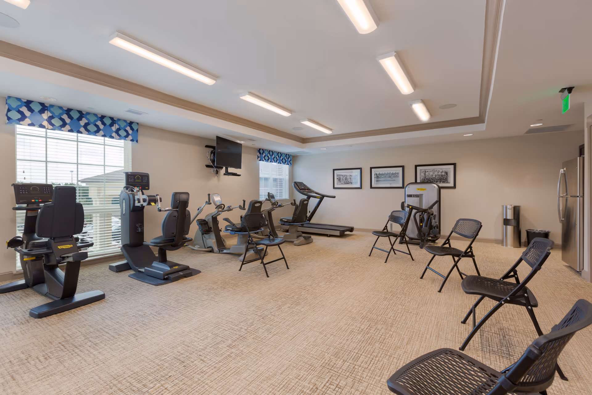 A senior living facility exercise room with several pieces of cardio equipment including recumbent bikes, upright bikes, and a treadmill. There are multiple black folding chairs arranged around the room, beige carpet, large windows with blue patterned valances, and framed black and white pictures on the wall. A flat screen TV is mounted on the wall.