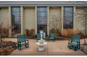 Outdoor patio area with four green wooden chairs arranged around a white decorative fountain. The patio is paved and surrounded by landscaping with dried plants and bushes. The background features large windows set in a beige building wall.