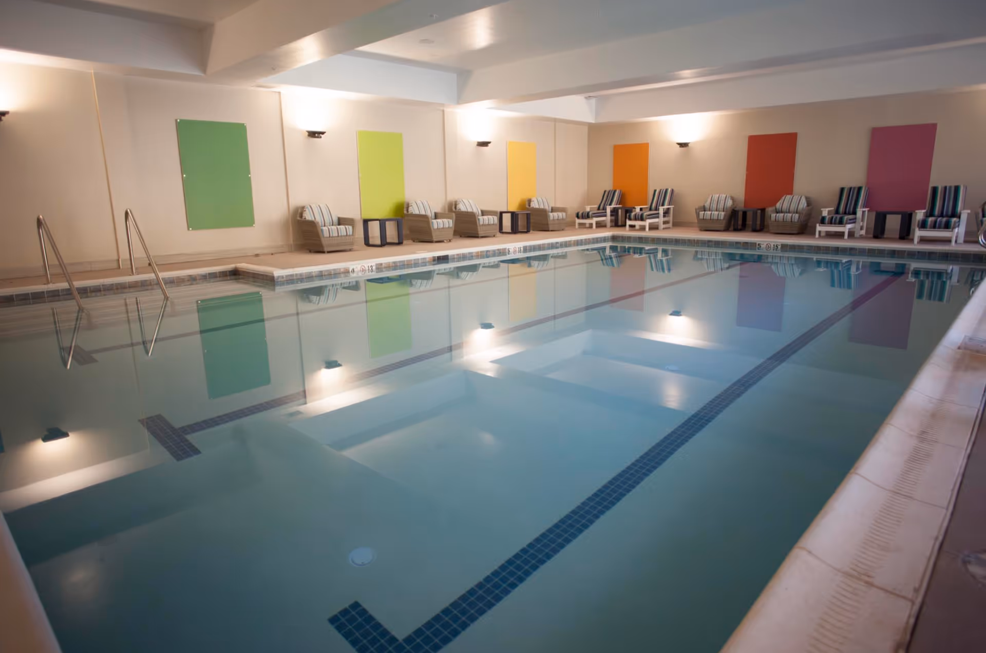 Indoor swimming pool with clear water, surrounded by a tiled deck. Along one wall, there are several cushioned lounge chairs and small tables, with colorful rectangular wall decorations in green, yellow, orange, red, and purple. The pool area is well-lit with wall-mounted lights.