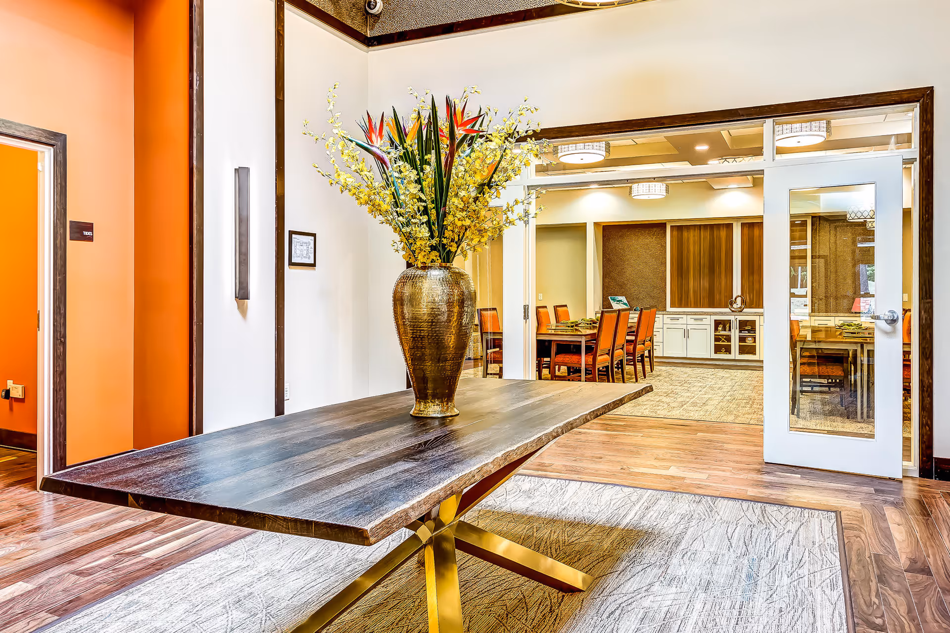 A modern interior space featuring a wooden table with a large decorative vase filled with yellow and red flowers. The room has wooden flooring and a light-colored rug underneath the table. In the background, there is a dining area with a long table and several orange chairs, visible through a glass door.