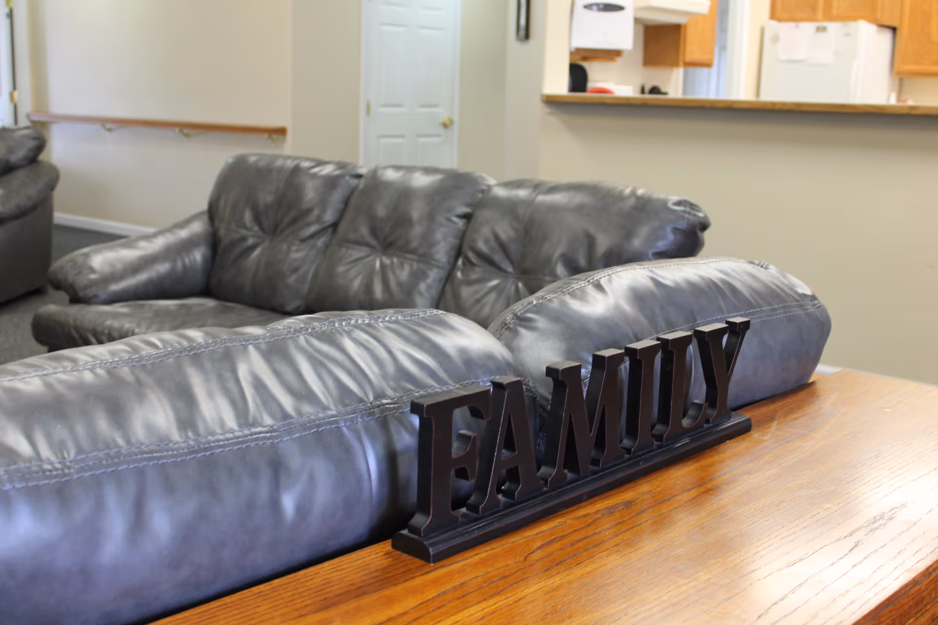 Interior view of a living room area with black leather sofas and a wooden table behind one sofa displaying a decorative black sign that reads 'FAMILY'. In the background, there is a partial view of a kitchen counter and a closed white door.