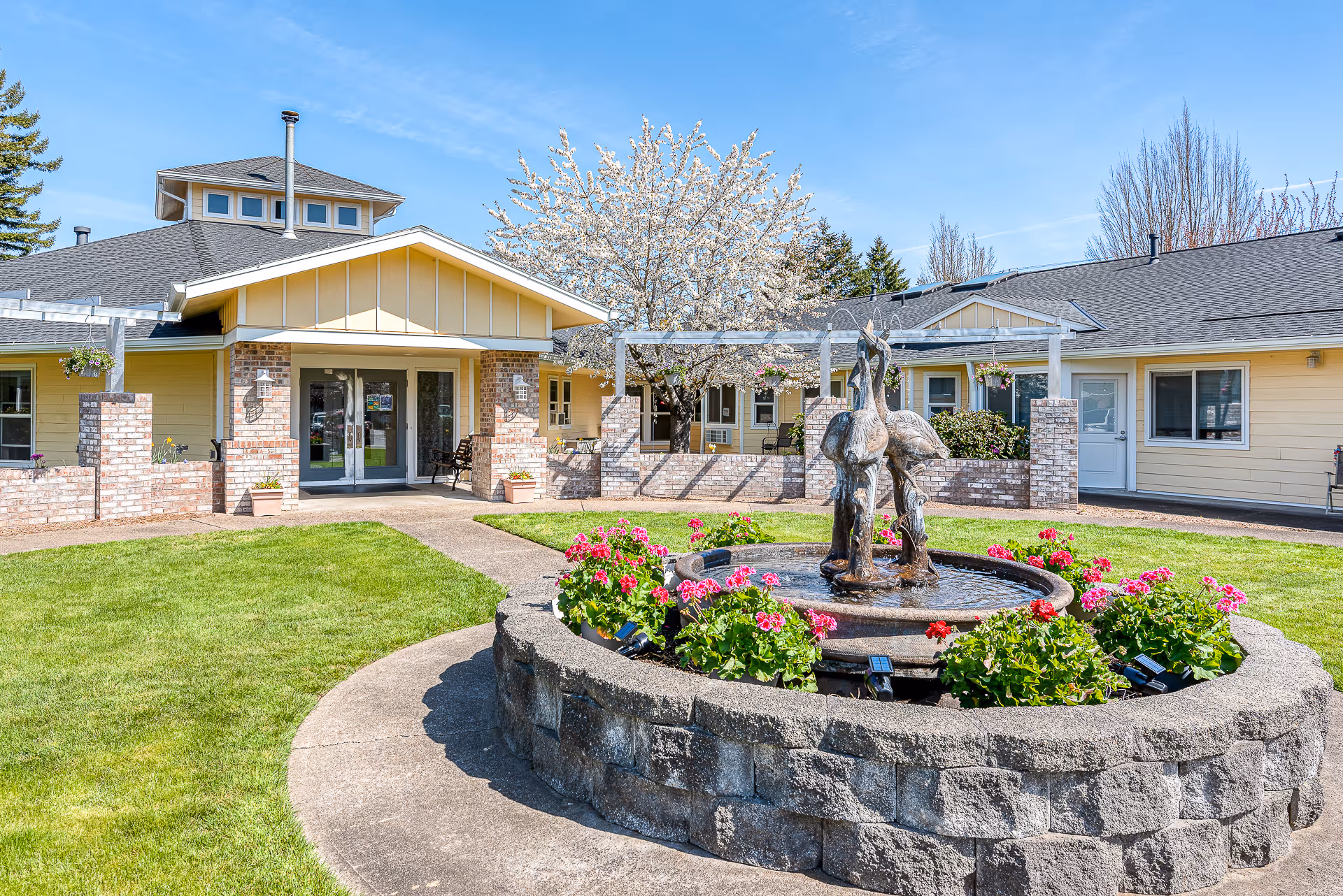 Outdoor courtyard area of Forest Grove Beehive Assisted Living featuring a circular stone fountain with bird sculptures surrounded by pink flowers, a well-maintained lawn, and a yellow building with brick accents and a covered entrance under a clear blue sky.