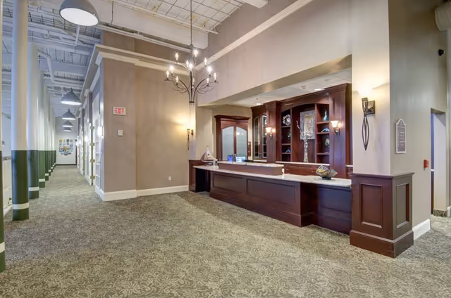 Interior view of a senior living facility hallway with a long carpeted corridor on the left and a wooden reception desk with decorative shelves and lighting on the right. The ceiling is high with exposed beams and hanging light fixtures, and the walls are painted in neutral tones.