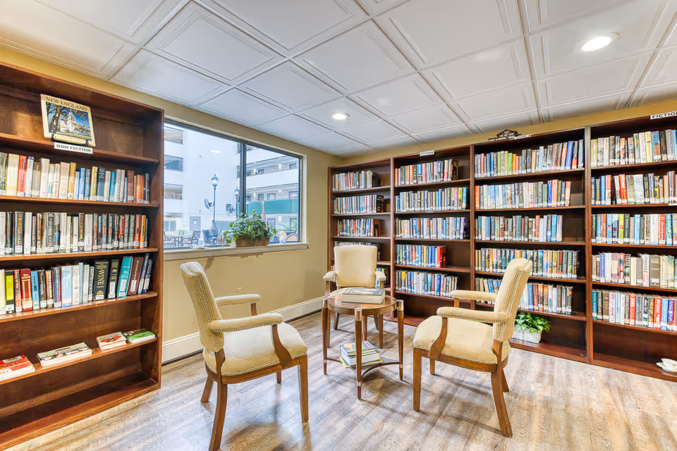 A cozy library room with wooden bookshelves filled with books labeled Non Fiction, Fiction, and Mystery. There are three upholstered armchairs arranged around a small wooden table with books on it. A window lets in natural light and shows an outdoor area with a lamp post and building exterior.