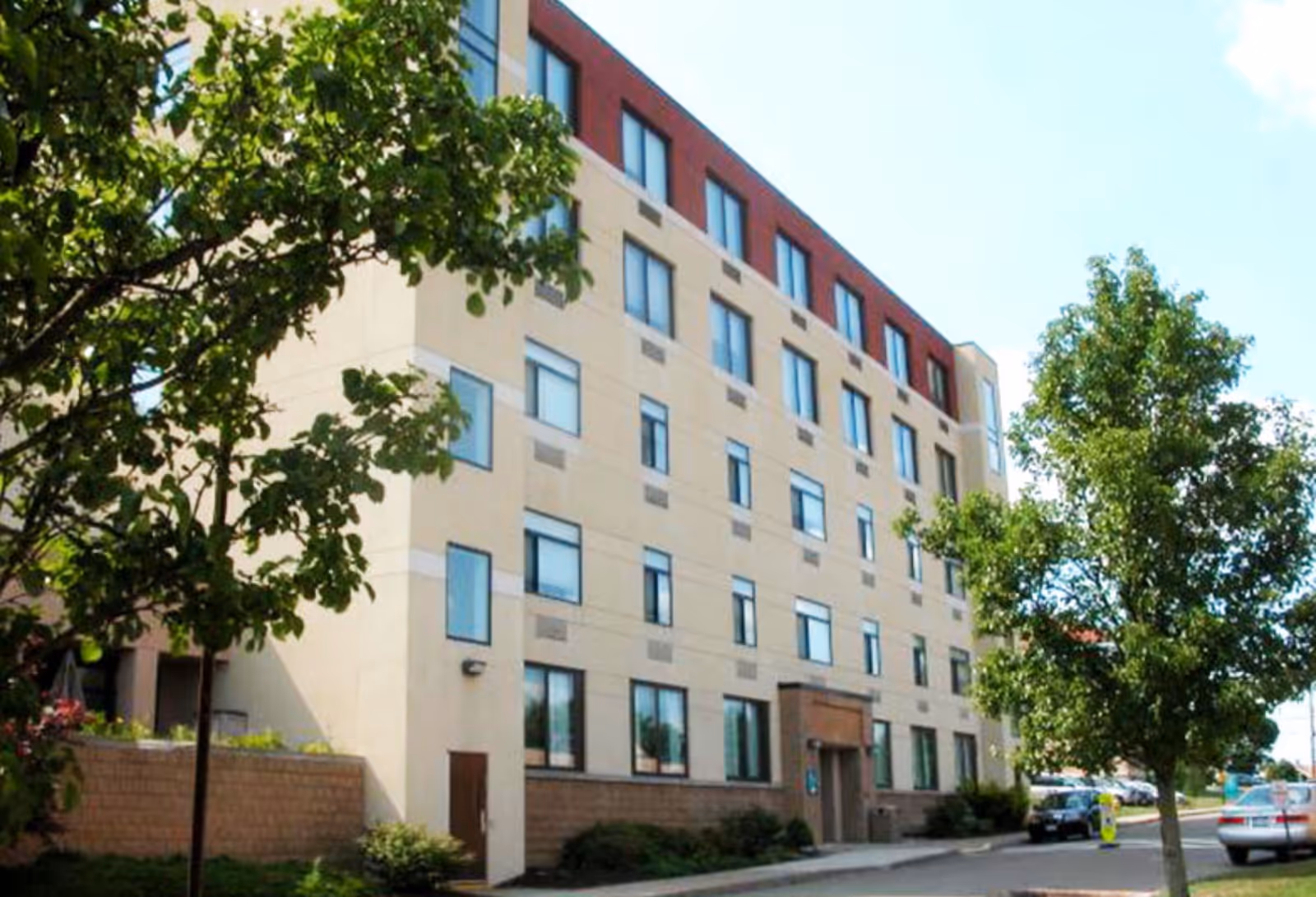 Exterior view of a multi-story beige and red senior living building with trees and a sidewalk.