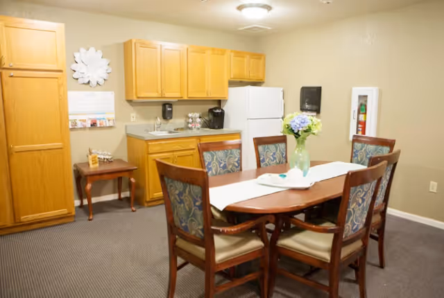 A small kitchen and dining area in a senior living facility featuring wooden cabinets, a white refrigerator, a sink, and a coffee maker. In front of the kitchen is a wooden dining table with six cushioned chairs, a white table runner, and a vase with flowers. The walls are beige, and the floor is carpeted.