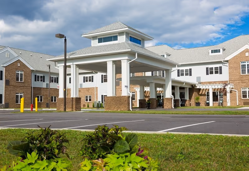 Exterior view of The Crossings At Blacksburg senior living facility showing a covered entrance with white columns and brick bases, surrounded by a parking lot and green landscaping under a partly cloudy sky.