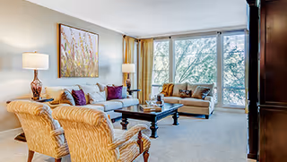 Bright living room with large windows, beige sofas and armchairs around a central coffee table and artwork on the wall.