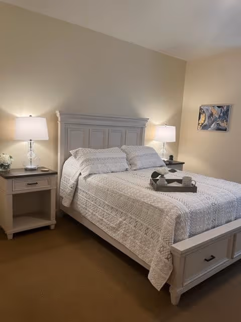 A neatly made bed with a white quilted bedspread and two pillows in a bedroom. There are two matching nightstands on either side of the bed, each with a lamp and some decorative items. A small abstract painting is hung on the wall to the right.