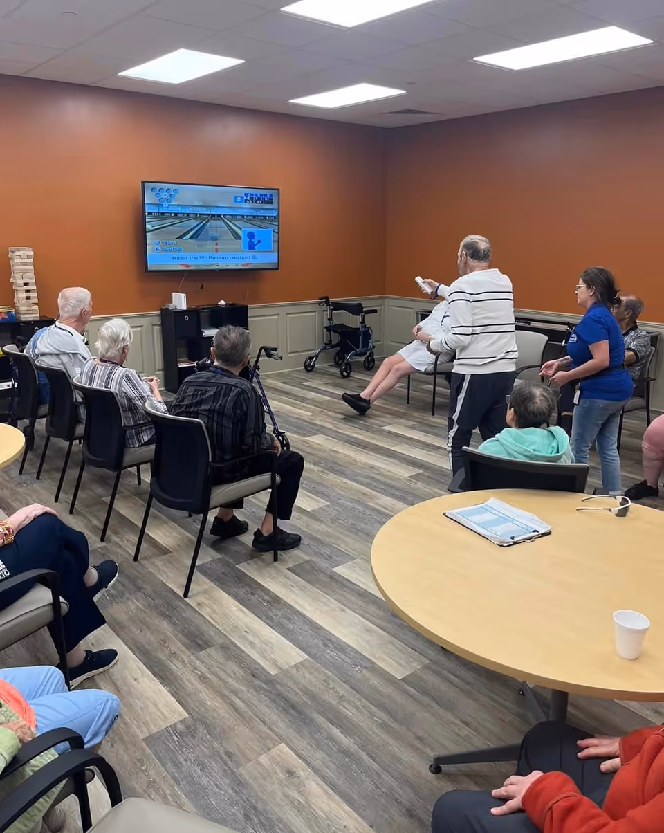 A group of elderly people seated and standing in a common room with wood-patterned flooring and orange walls, watching and playing a bowling game on a wall-mounted TV. Some individuals are using walkers and a staff member is assisting one of the players.