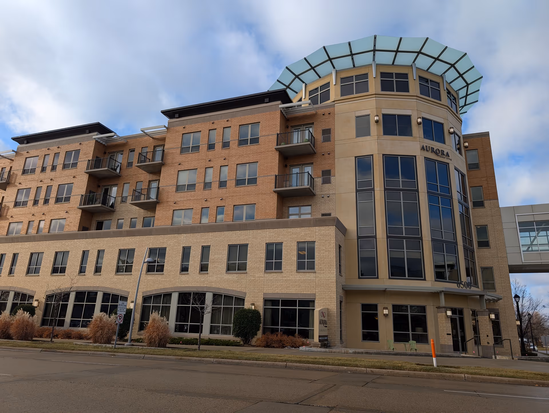 Exterior view of a multi-story senior living facility named Aurora on France, featuring large windows, balconies, and a distinctive rounded corner with a glass canopy on top. The building is made of light-colored bricks and has a sidewalk and street in front.