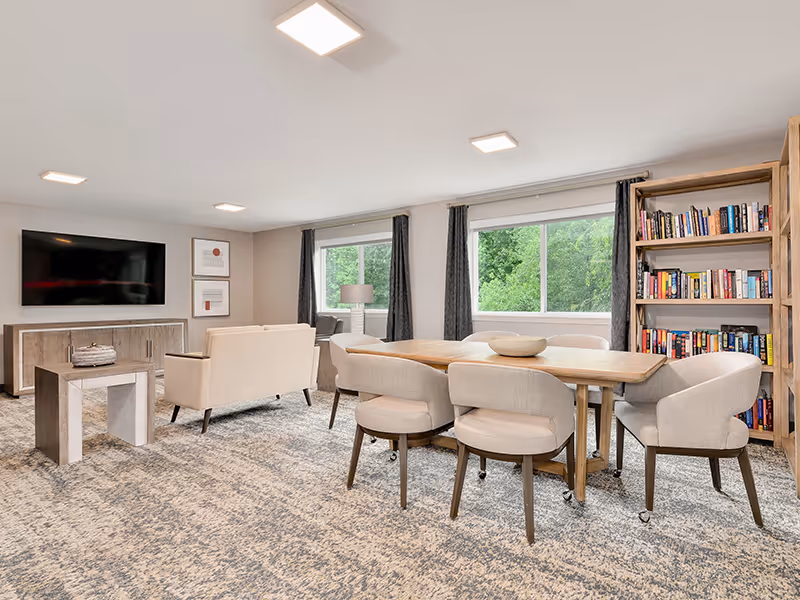 A bright and spacious senior living community common area featuring a wooden dining table with six upholstered chairs on wheels, a beige loveseat and armchair facing a wall-mounted flat screen TV, a wooden bookshelf filled with books, two large windows with dark curtains letting in natural light, and a patterned carpet covering the floor.