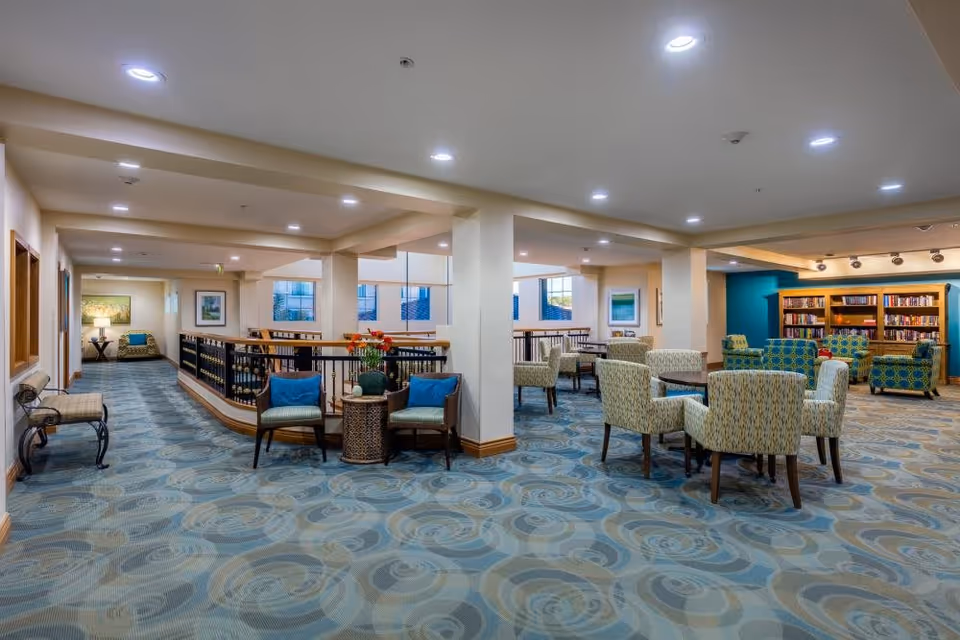 A spacious, well-lit common area in a senior living facility featuring multiple seating arrangements with cushioned chairs and small tables. The room has patterned blue and beige carpeting, light-colored walls, and several framed artworks. In the background, there is a bookshelf filled with books against a teal accent wall. The area appears comfortable and inviting for socializing or reading.