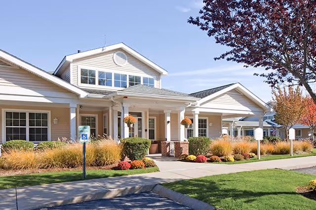 Exterior view of The Cottages of Dartmouth Village showing a single-story building with beige siding, white trim, and a covered entrance supported by white columns. The landscaping includes bushes, ornamental grasses, and colorful flowers, with a sidewalk and a handicapped parking sign visible in front.