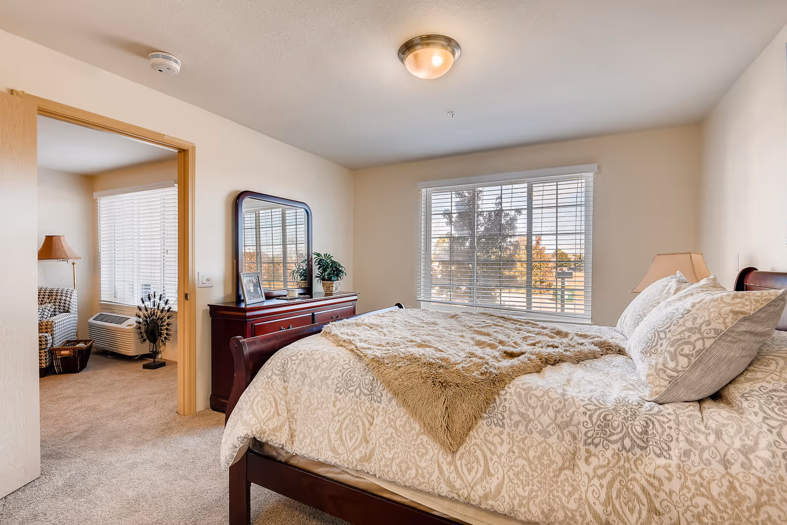 A bright bedroom with a large window covered by white blinds, a bed with patterned bedding and a beige throw blanket, a wooden dresser with a mirror, a small plant, and a photo frame. An open door leads to an adjacent room with a window, a patterned armchair, a floor lamp, and an air conditioning unit.