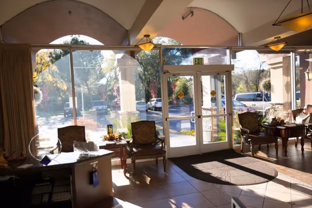 Interior view of a senior living facility lobby with large glass doors and windows letting in sunlight. There are several wooden chairs with cushions and small tables arranged near the entrance. Outside, cars are parked and trees are visible.