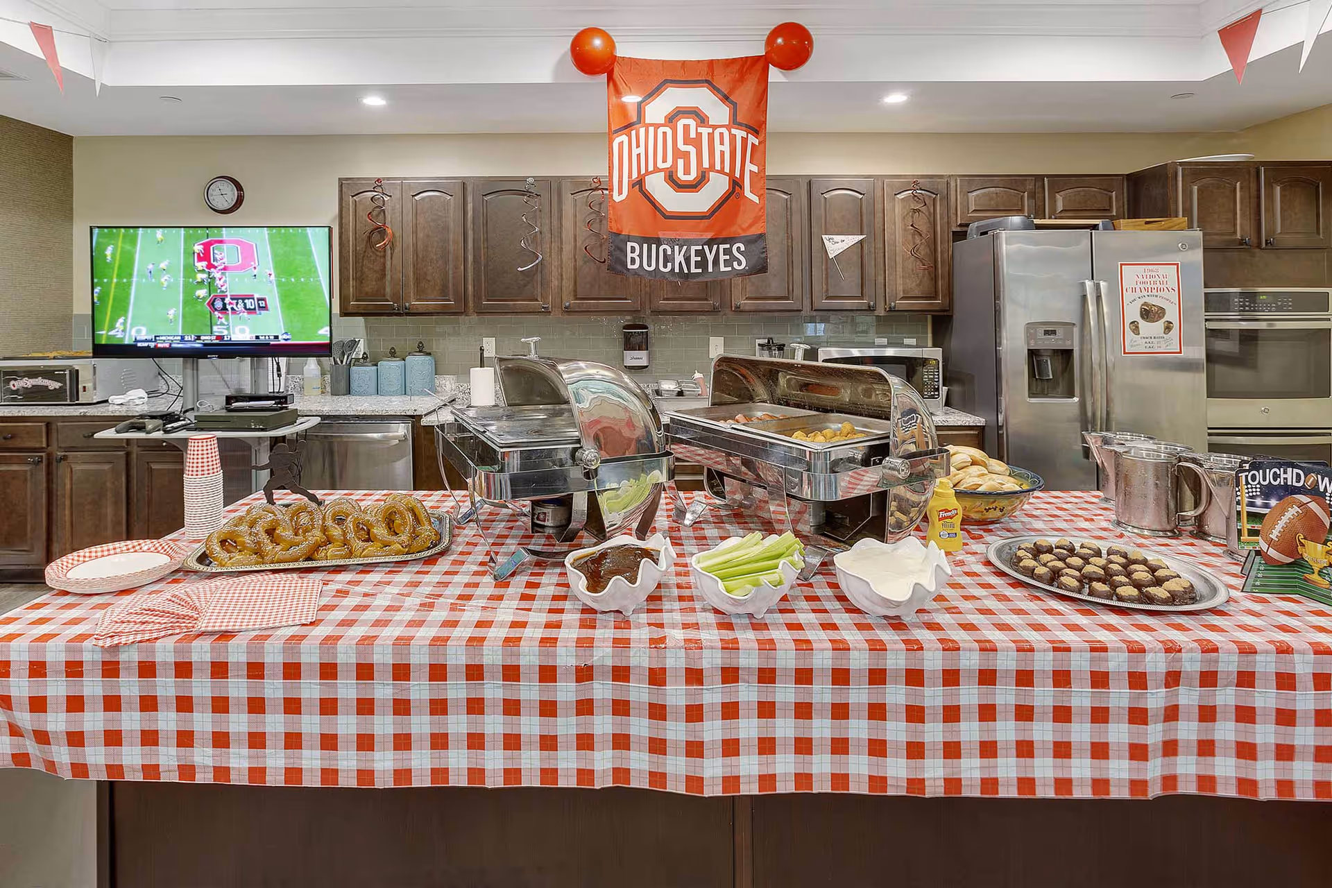 A kitchen area decorated with Ohio State Buckeyes themed items including a hanging flag and a TV showing a football game. A table covered with a red and white checkered tablecloth holds trays of food such as pretzels, celery sticks, sauces, rolls, and dessert balls. Stainless steel appliances and wooden cabinets are visible in the background.