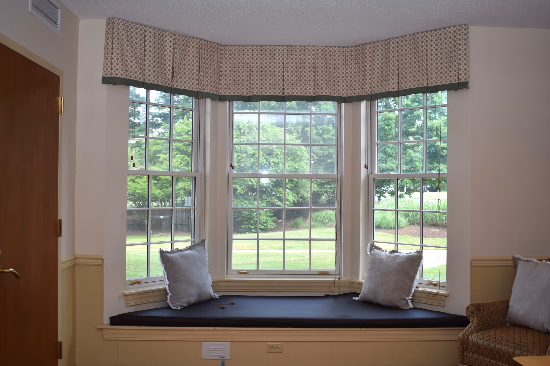 A cozy window seat with a black cushion and two light gray pillows set in a bay window with three large panes. The window has a patterned valance at the top, and outside greenery is visible through the glass. To the right, part of a cushioned armchair with a similar gray pillow is visible, and to the left is a closed wooden door with a brass handle.
