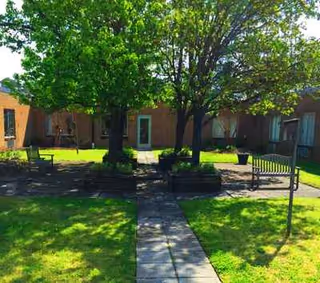 Sunlit courtyard with green lawns, trees, benches, and a concrete path between brick buildings.