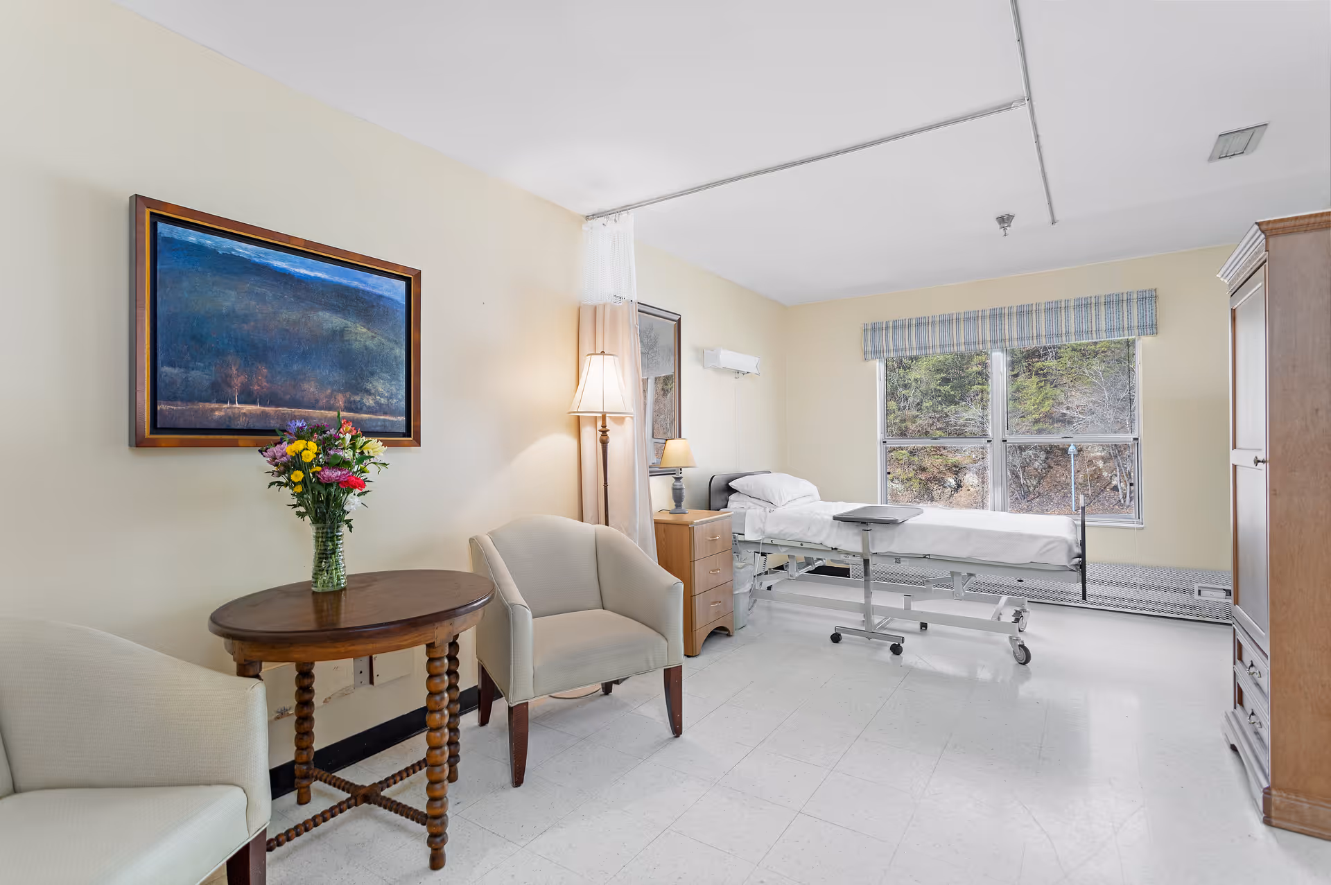 A bright and clean patient room with a hospital bed positioned near a window showing an outdoor view. The room includes two beige armchairs around a small wooden table with a vase of colorful flowers, a wooden nightstand with a lamp, a framed landscape painting on the wall, and a wooden wardrobe.