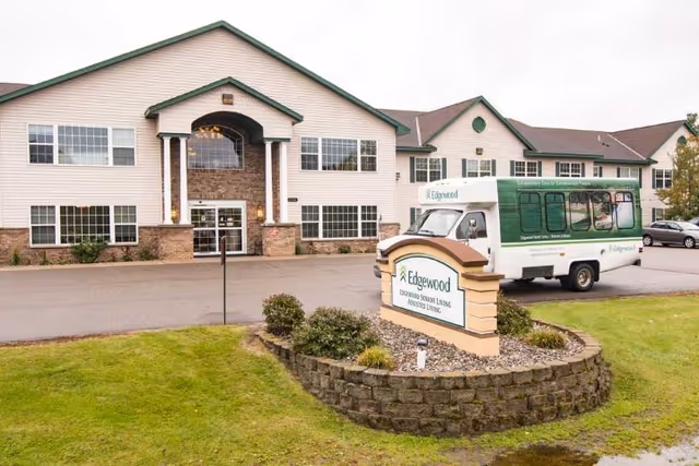 Exterior view of Edgewood Baxter, a two-story senior living facility with beige siding and stone accents. There is a green and white shuttle bus parked near the entrance, and a landscaped sign in front displaying the facility's name. The area is surrounded by green grass and a paved driveway.