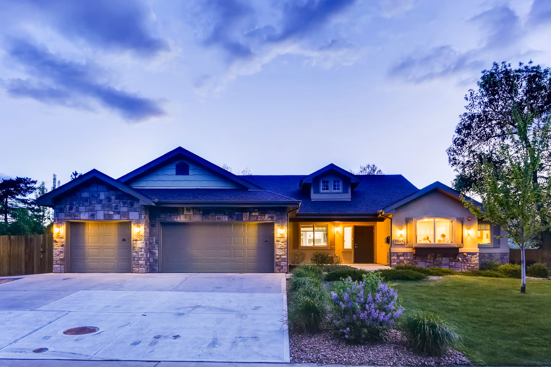 Exterior front view of a single-story residential-style building with a three-car garage, illuminated by outdoor lights at dusk. The building has stone and siding facade with a well-maintained lawn, shrubs, and a tree in the front yard under a partly cloudy sky.