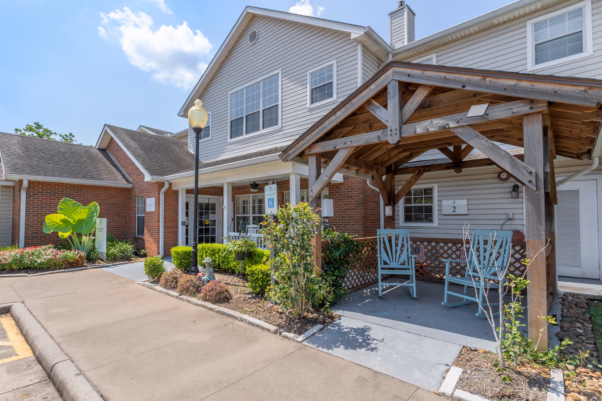 Exterior view of Brookdale The Heights facility showing a covered porch area with two blue rocking chairs, a wooden pergola, and a landscaped garden with bushes and flowers. The building has a combination of brick and siding with multiple windows and a street lamp near the entrance.