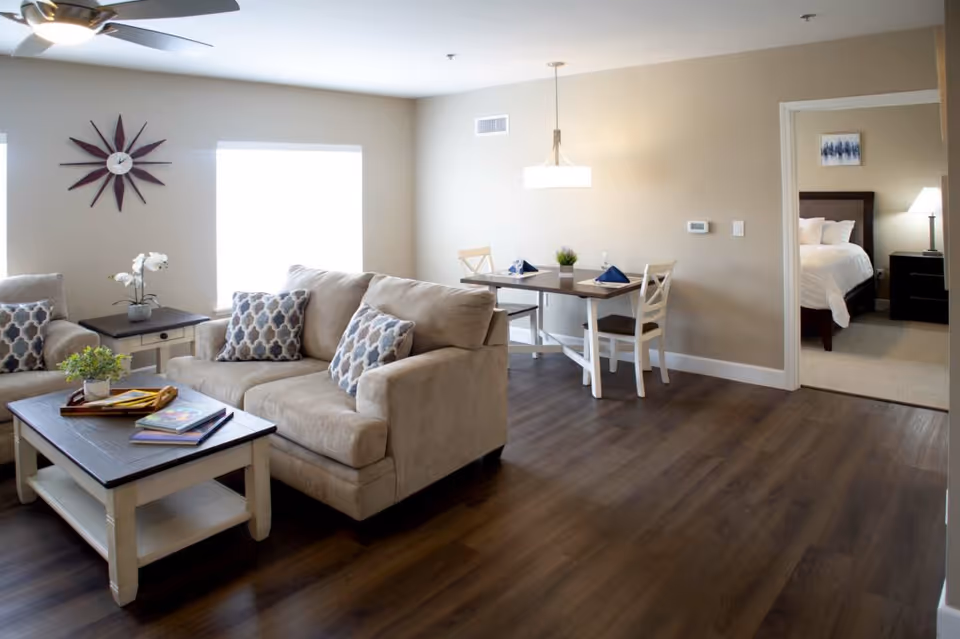 A bright and modern living room with beige sofas adorned with patterned cushions, a wooden coffee table with books and a small plant, and a side table with a white orchid. Adjacent to the living room is a dining area with a table set for two, featuring blue napkins and a small plant centerpiece. In the background, an open doorway reveals a bedroom with a neatly made bed, a nightstand, and a lamp. The room has wooden flooring and neutral-colored walls.
