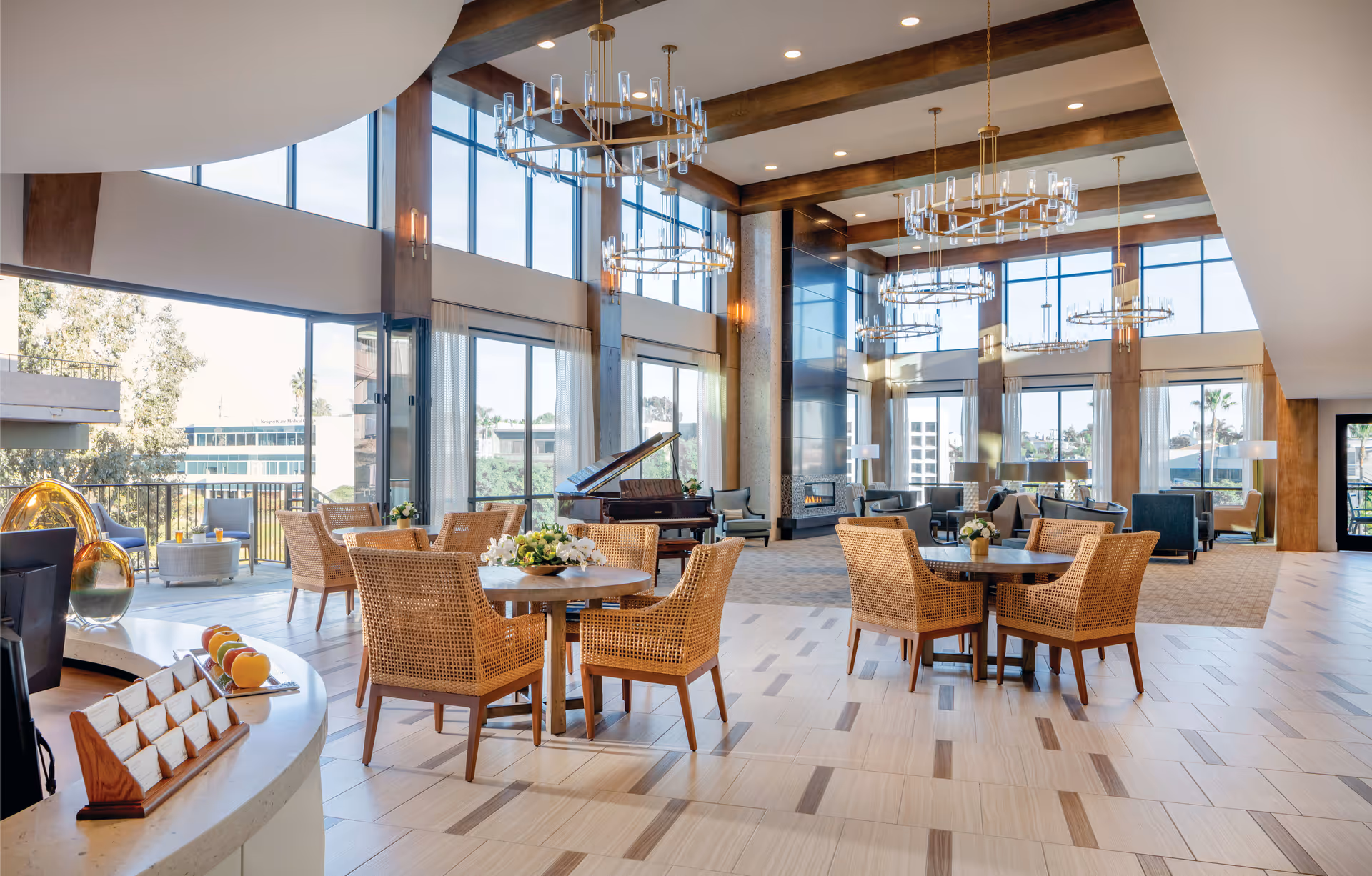 Spacious and bright senior living facility common area with large windows, wooden beams, modern chandeliers, wicker chairs around round tables, a grand piano, and a fireplace. The room has a mix of tiled and carpeted flooring with comfortable seating areas and decorative plants.