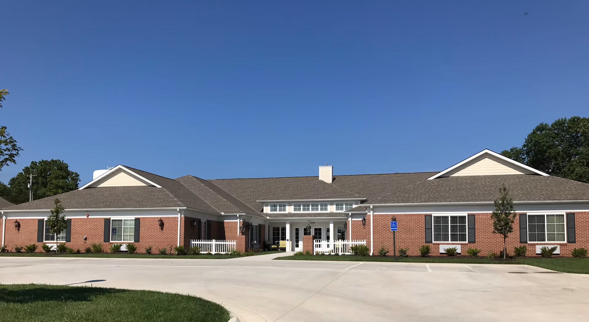 Front exterior view of a single-story brick building with a gray shingled roof, white trim, and black shutters. The building has a covered entrance with white columns and a small white picket fence on either side. There are two handicap parking signs in front of the building, and the sky is clear and blue.