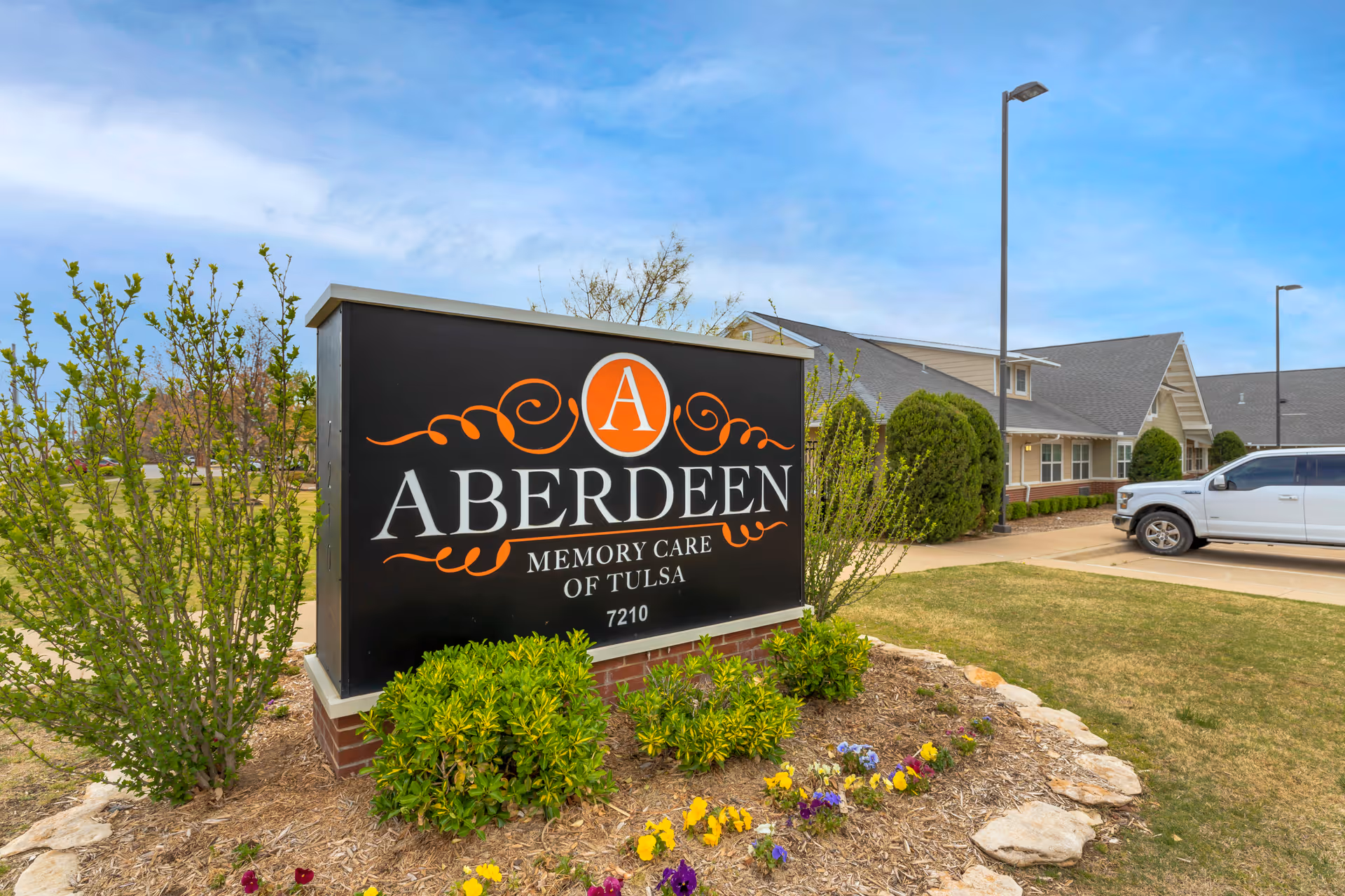 Outdoor view of the Aberdeen Memory Care of Tulsa facility sign surrounded by landscaping with bushes and flowers, with the building and a parked white vehicle visible in the background under a partly cloudy sky.