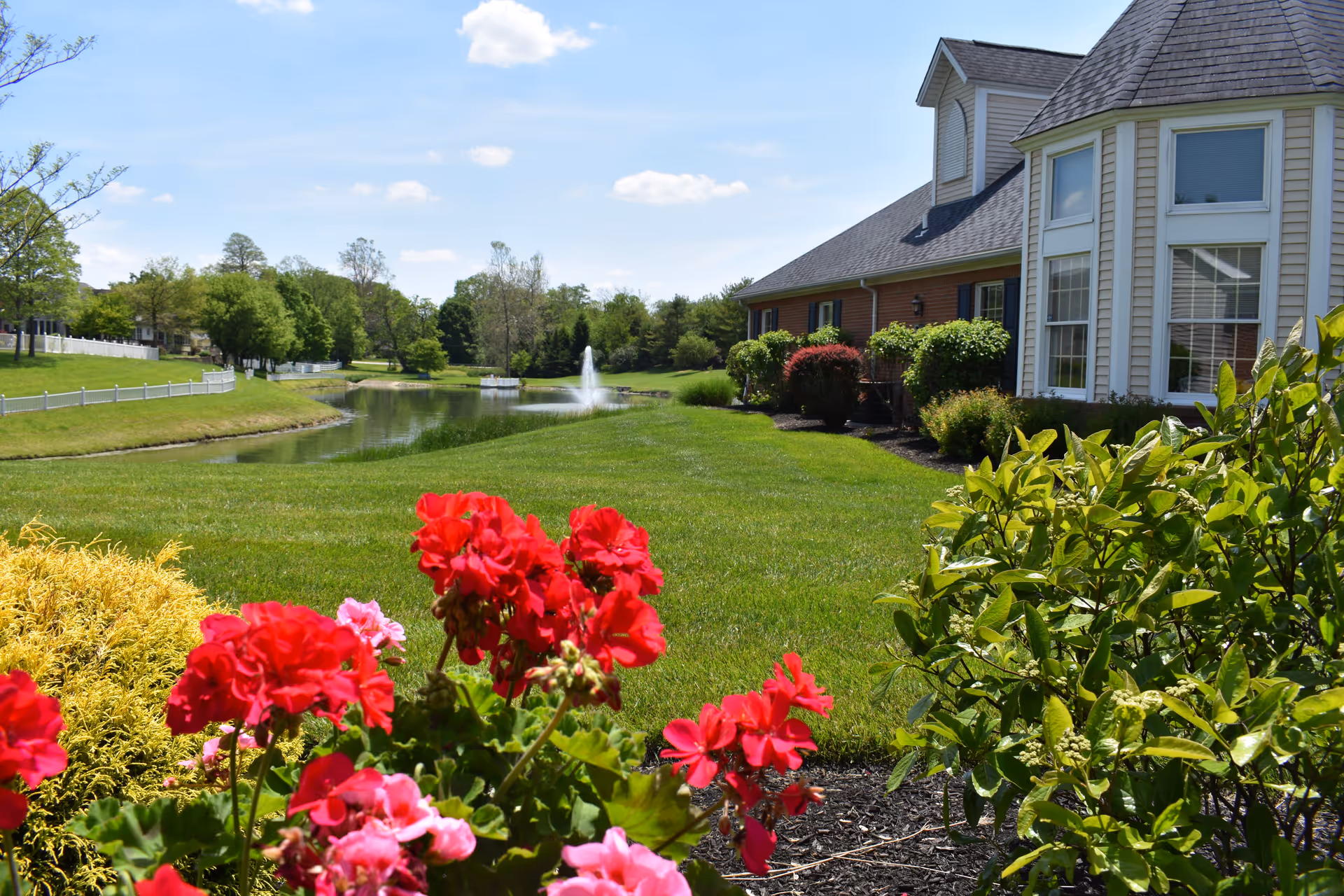 A scenic outdoor view at Ohio Living Cape May featuring a well-maintained lawn with vibrant red and pink flowers in the foreground, a small pond with a water fountain in the middle distance, and a building with large windows and beige siding on the right side. Trees and a clear blue sky with a few clouds are visible in the background.