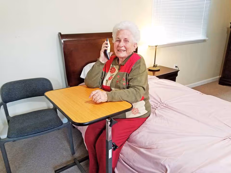An elderly woman with white hair is sitting on a bed in a bedroom, smiling and holding a phone to her ear. She is wearing a green and red sweater and red pants. There is a small overbed table in front of her and a gray chair beside it. The room has a wooden headboard, a nightstand with a lamp, and a window with blinds.