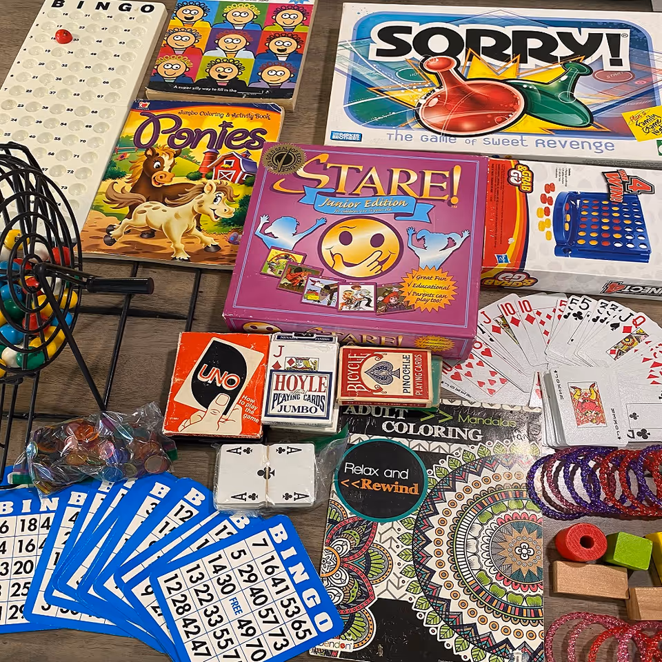 Tabletop assortment of games and activity supplies including Bingo cards, UNO, Sorry!, playing cards, a Stare! box, and coloring books.