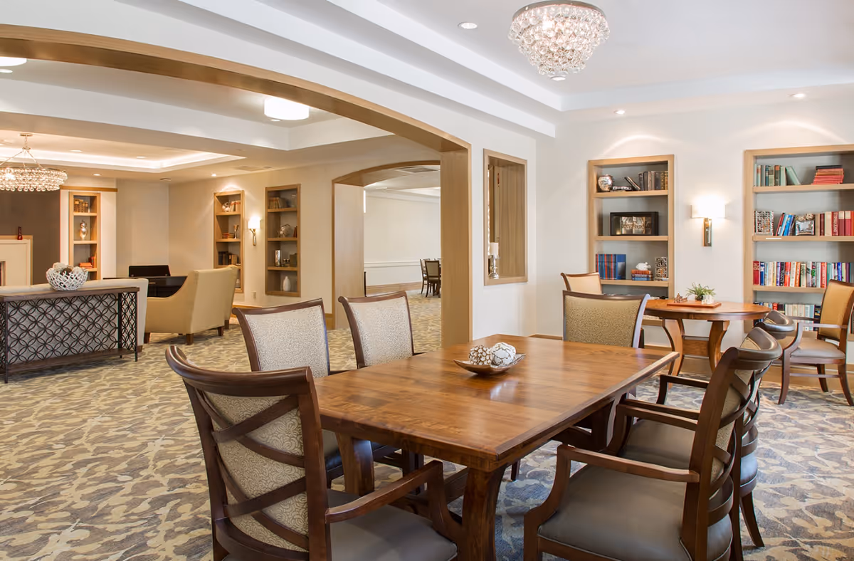Well-lit communal dining and lounge area with a large wooden table, upholstered chairs, bookshelves, and chandeliers.