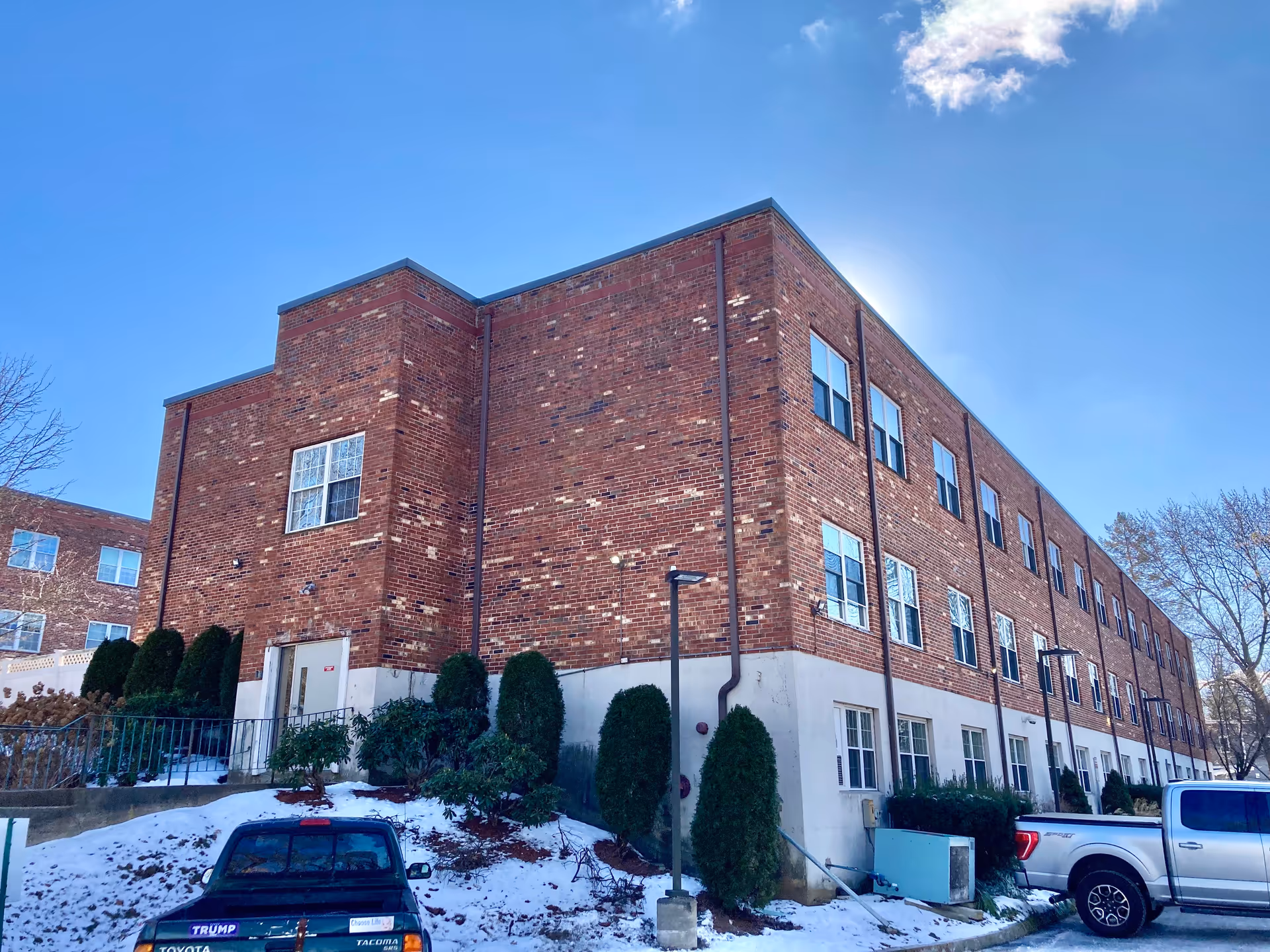 Exterior view of a three-story brick building with multiple windows, surrounded by snow-covered ground, bushes, and parked vehicles under a clear blue sky.