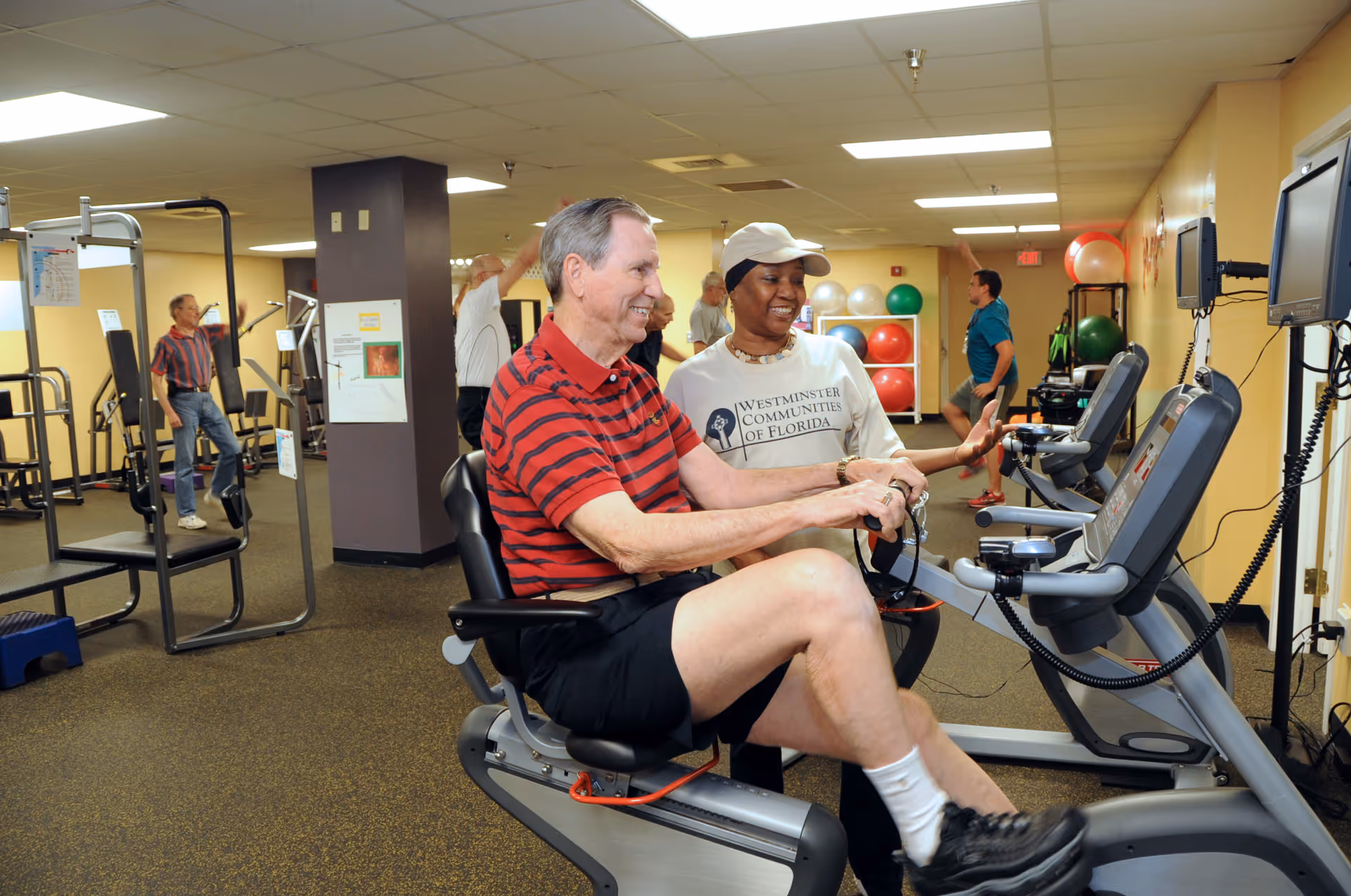 An elderly man in a red and black striped shirt exercises on a recumbent stationary bike in a fitness room. A woman wearing a Westminster Communities of Florida shirt and a white cap is smiling and assisting him. In the background, other seniors are using various exercise equipment and stretching.