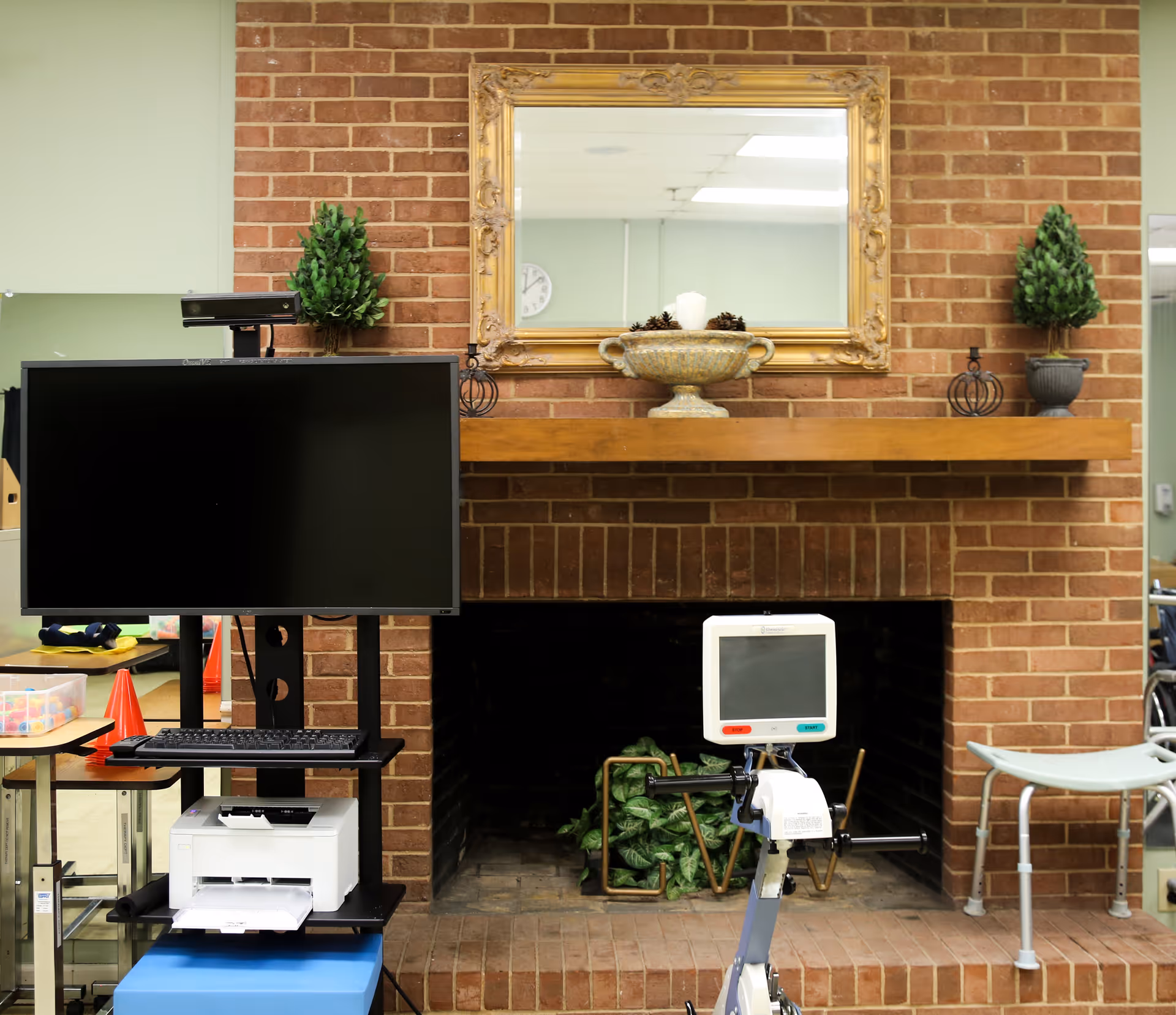 An indoor room with a brick fireplace and a wooden mantel decorated with a large ornate mirror, two small potted plants, and a decorative bowl with pinecones and a candle. In front of the fireplace, there is medical equipment including a monitor and a stationary exercise bike. To the left, there is a computer monitor on a stand with a keyboard and printer below it. The room has light green walls and fluorescent ceiling lights.