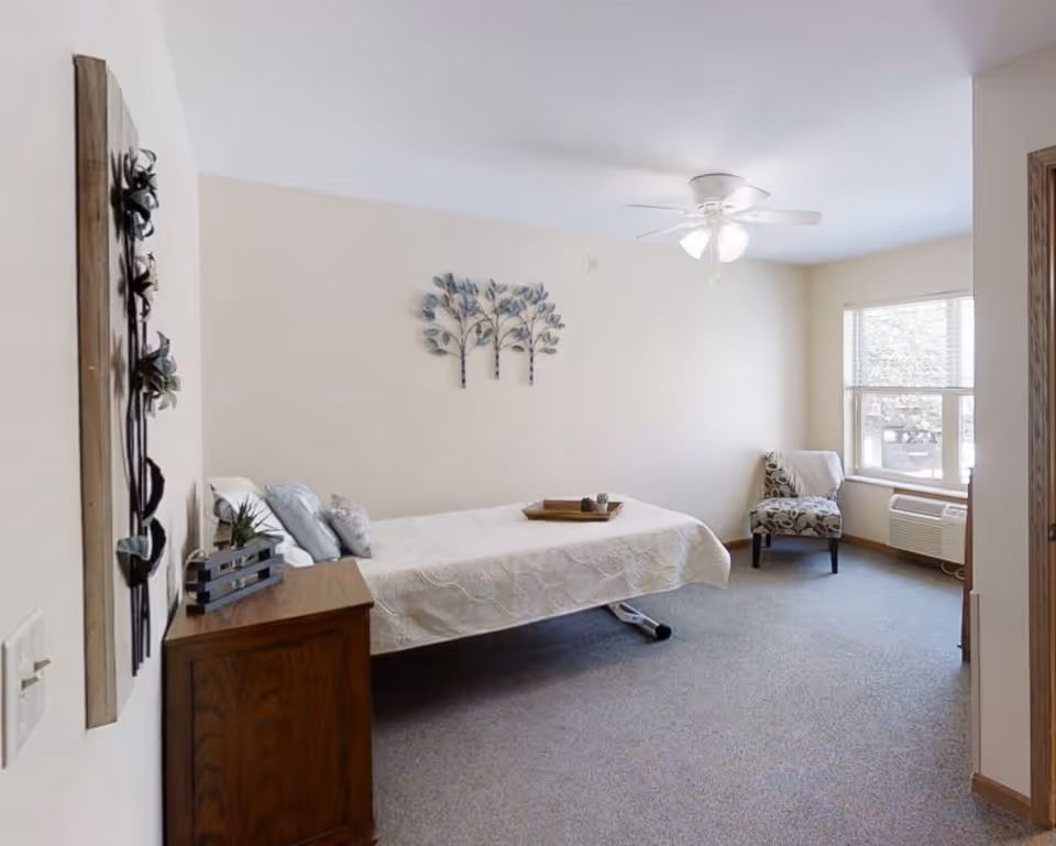 A simple, clean bedroom with a single bed covered in a white quilt, a wooden nightstand with decorative items, a patterned armchair near a window with blinds, a ceiling fan with lights, and neutral-colored walls adorned with metal tree wall art.