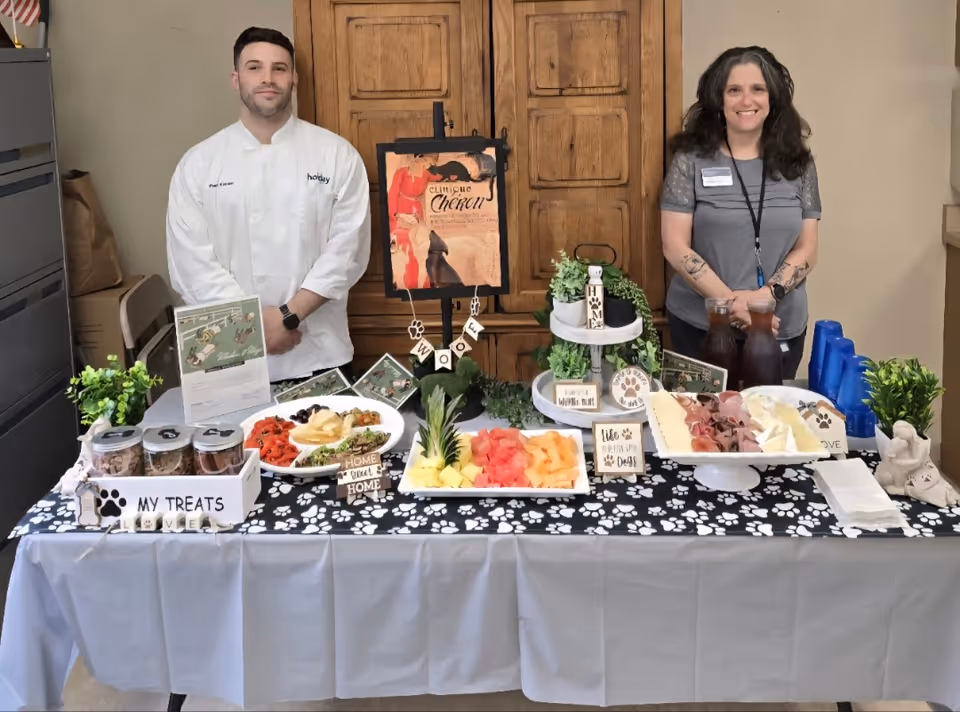 Two people standing behind a table covered with a white and black paw-print tablecloth. The table displays various food items including fruit, cheese, and meats, along with decorative signs and plants. The person on the left is wearing a white chef's coat, and the person on the right is wearing a gray shirt with a name badge. The background features wooden cabinets.