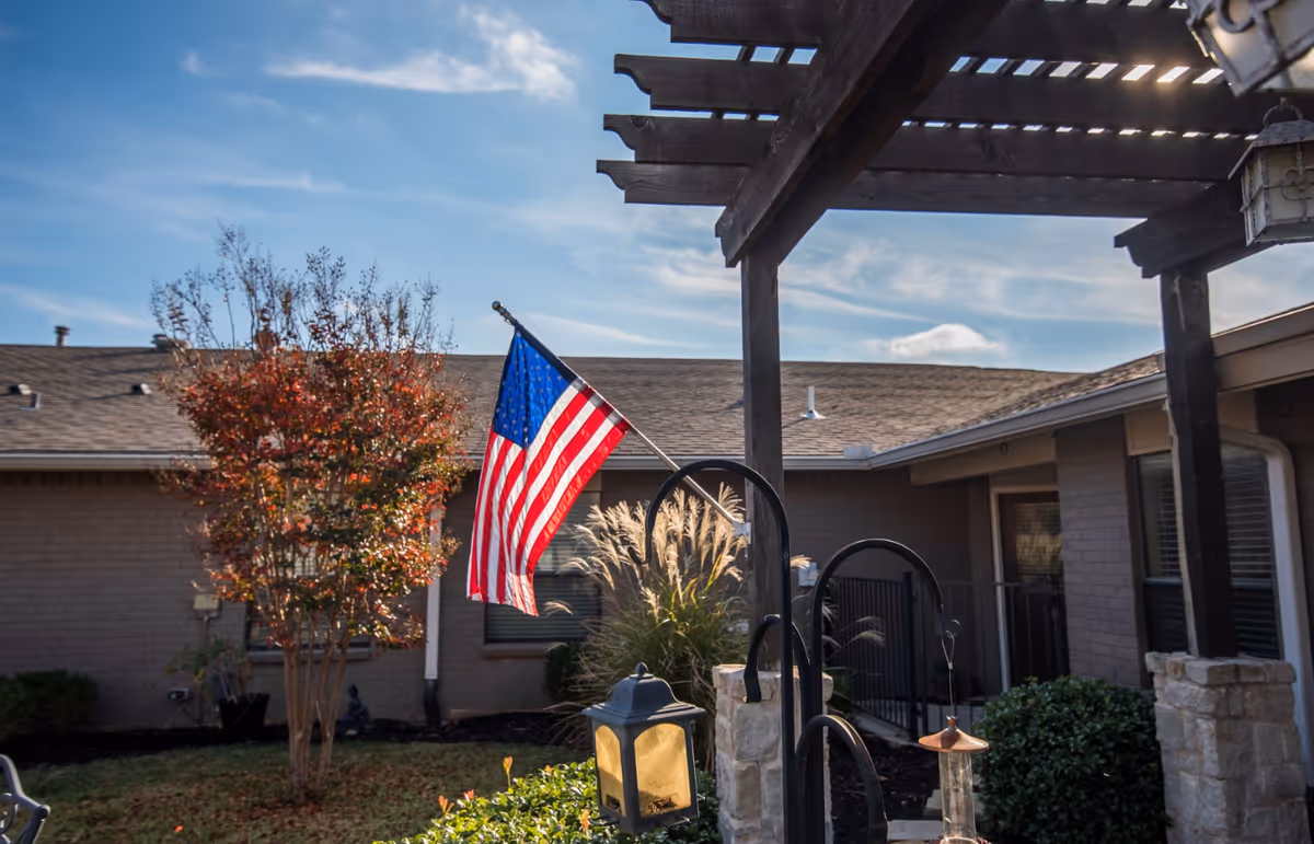 An American flag hangs from a pergola in a landscaped courtyard outside a single-story residential building.