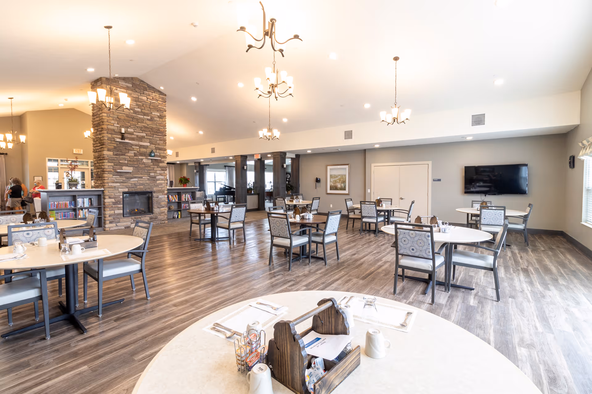 Spacious dining and common area with round tables and chairs, a stone fireplace, chandeliers, and a wall-mounted TV.